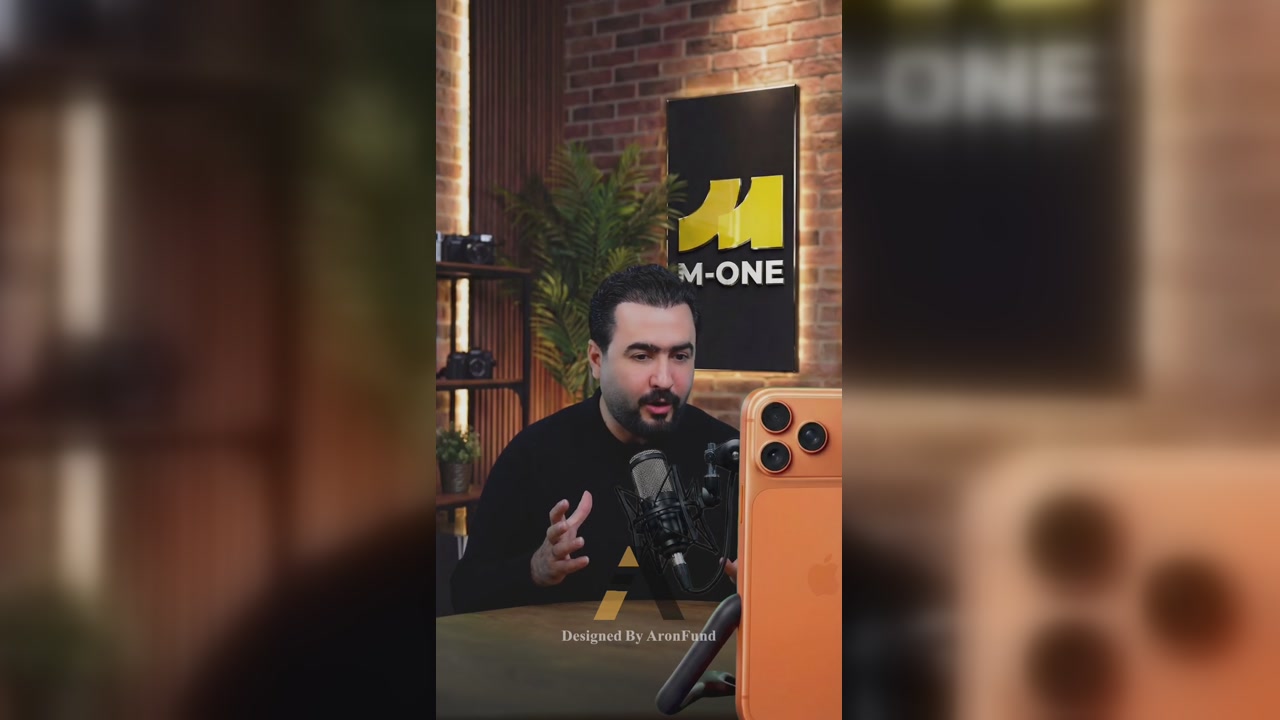 A presenter speaking at a studio microphone in an AI-generated brick-wall set, with an orange smartphone composited beside him